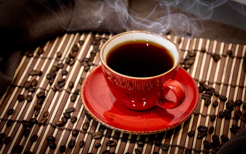A steaming cup of coffee in a red mug surrounded by coffee beans on a bamboo mat